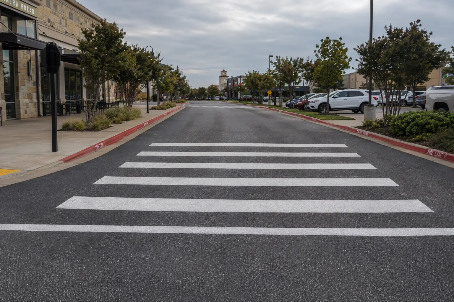 A finished thermoplastic crosswalk laid across a commercial drive lane — thick white bands, exactly parallel, evenly spaced, square to the kerbs at both ends, their surfaces catching the light with a faint glassy sparkle from the reflective beads
