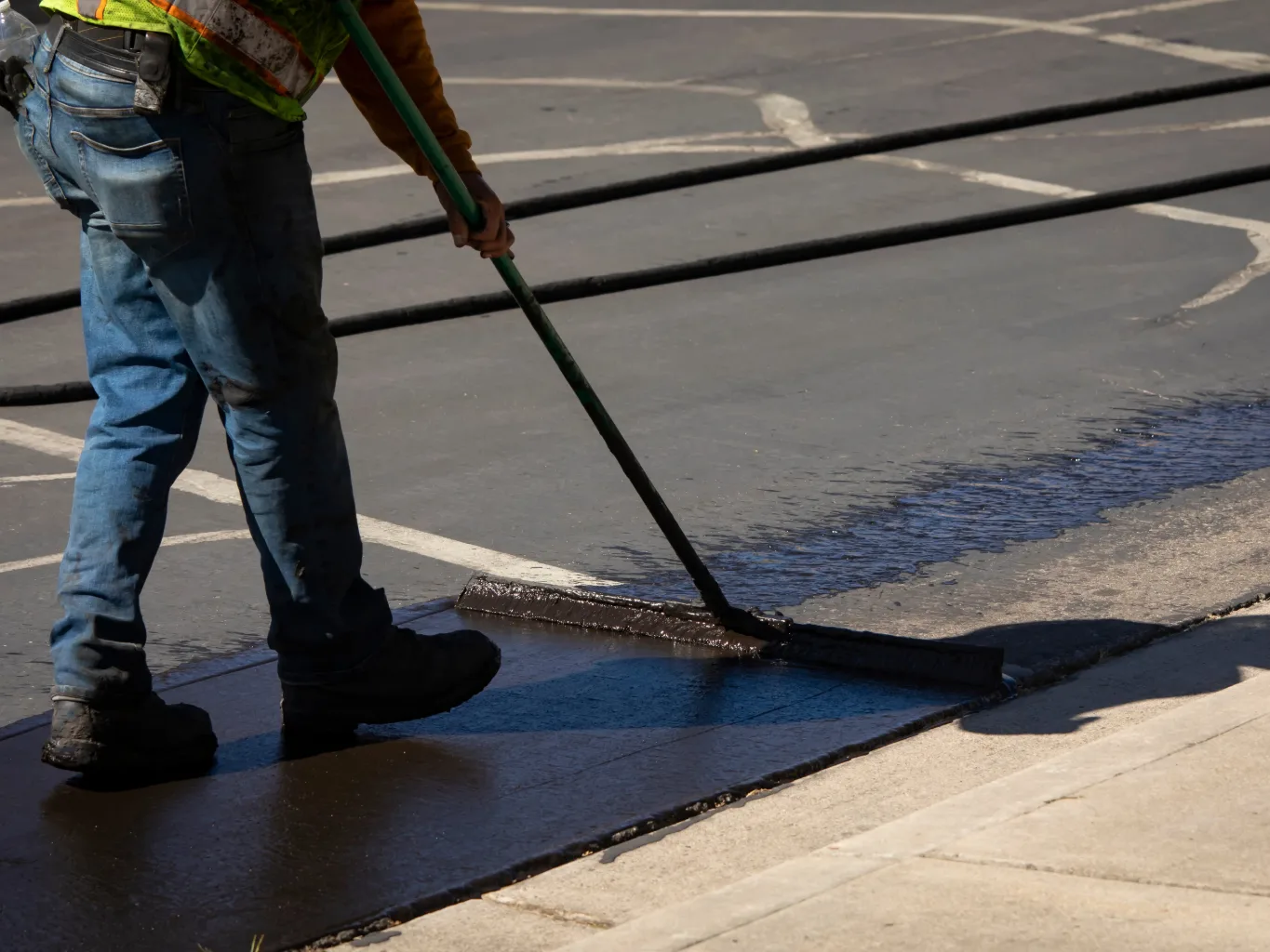Before any coating is applied, a striping contractor uses a backpack blower to clear loose dirt and debris from the edge of an asphalt parking area, focused on the pavement rather than the camera
