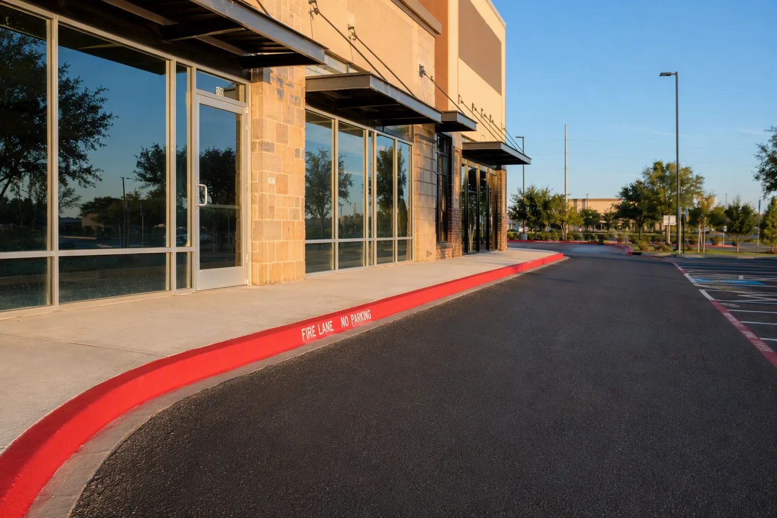 A long run of freshly coated bright red kerb curving gently along the front of a commercial building, the white words NO PARKING FIRE LANE stencilled clearly and correctly along it at even intervals
